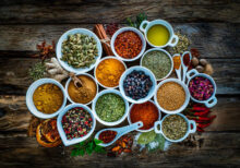 Top view of a rustic wood kitchen table filled with a large group of multi colored spices and herbs in white bowls. Spices and herb included are clove, turmeric, star anise, bay leaf, cinnamon, curry powder, ginger, nutmeg, peppercorns, cinnamon, salt, chili pepper, cardamom, dried oregano, basil, parsley, rosemary and dried orange slices. An olive oil bowl is included in the composition. High resolution 42Mp studio digital capture taken with SONY A7rII and Zeiss Batis 40mm F2.0 CF lens